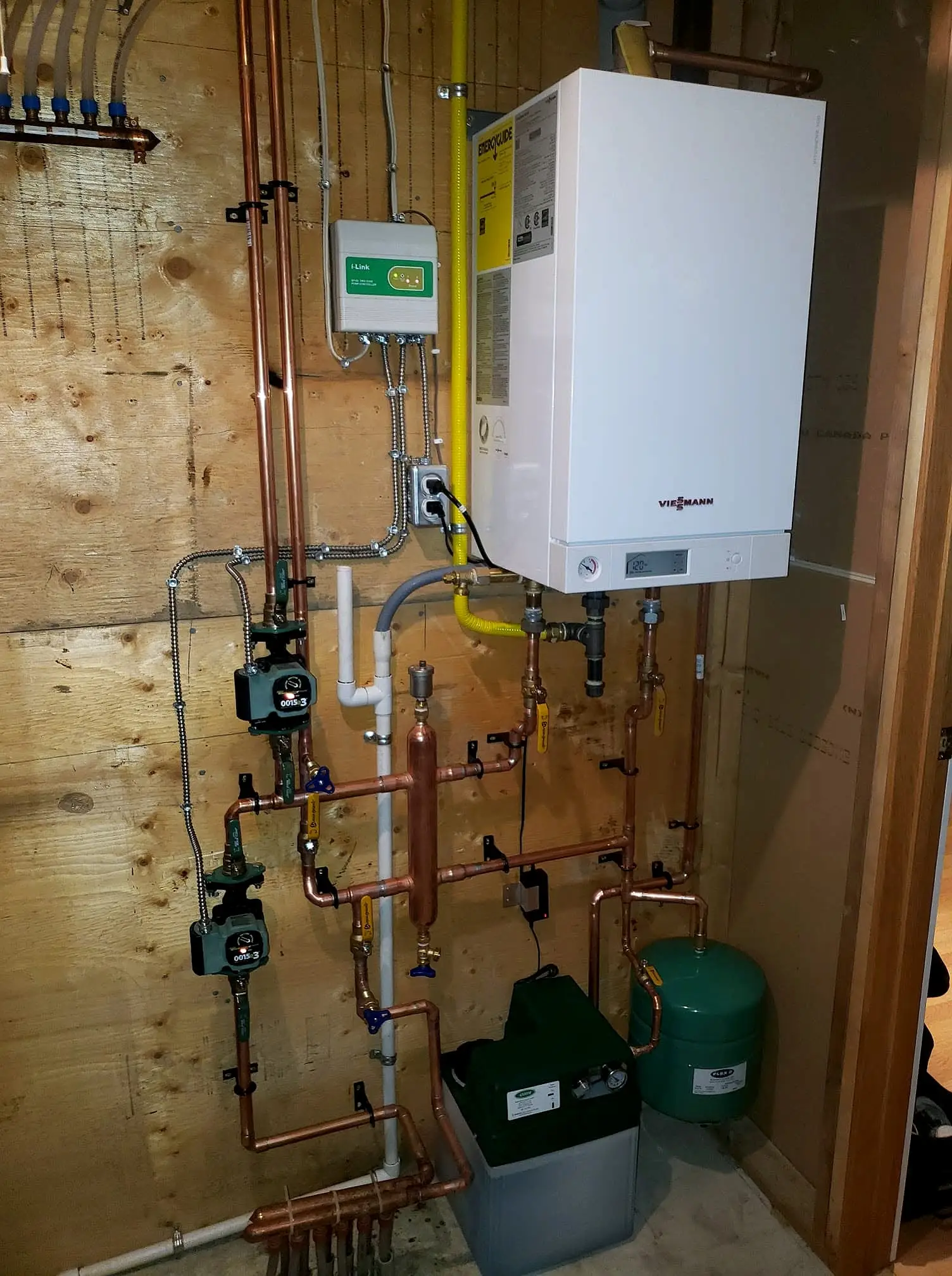 Wall-mounted high-efficiency boiler with copper near-boiler piping, circulator pumps and expansion tank on a plywood wall