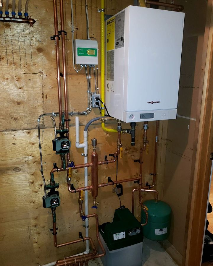 Wall-mounted high-efficiency boiler with copper near-boiler piping, circulator pumps and expansion tank on a plywood wall