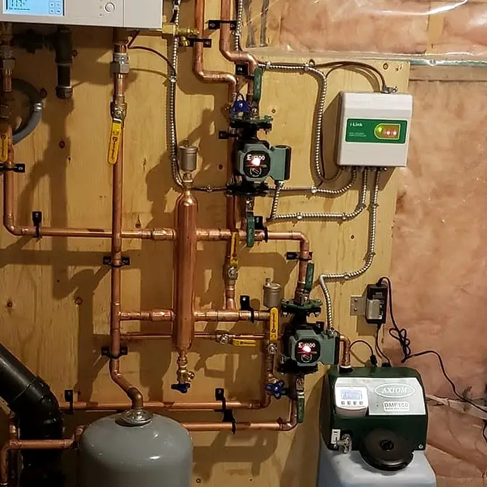 Close-up of copper piping with two circulator pumps and a pump control box below a wall-hung boiler