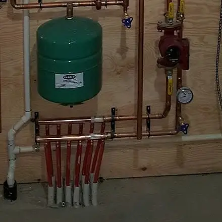 Red in-floor heating loops connected to a copper manifold, with an expansion tank, circulator pump and gauge above
